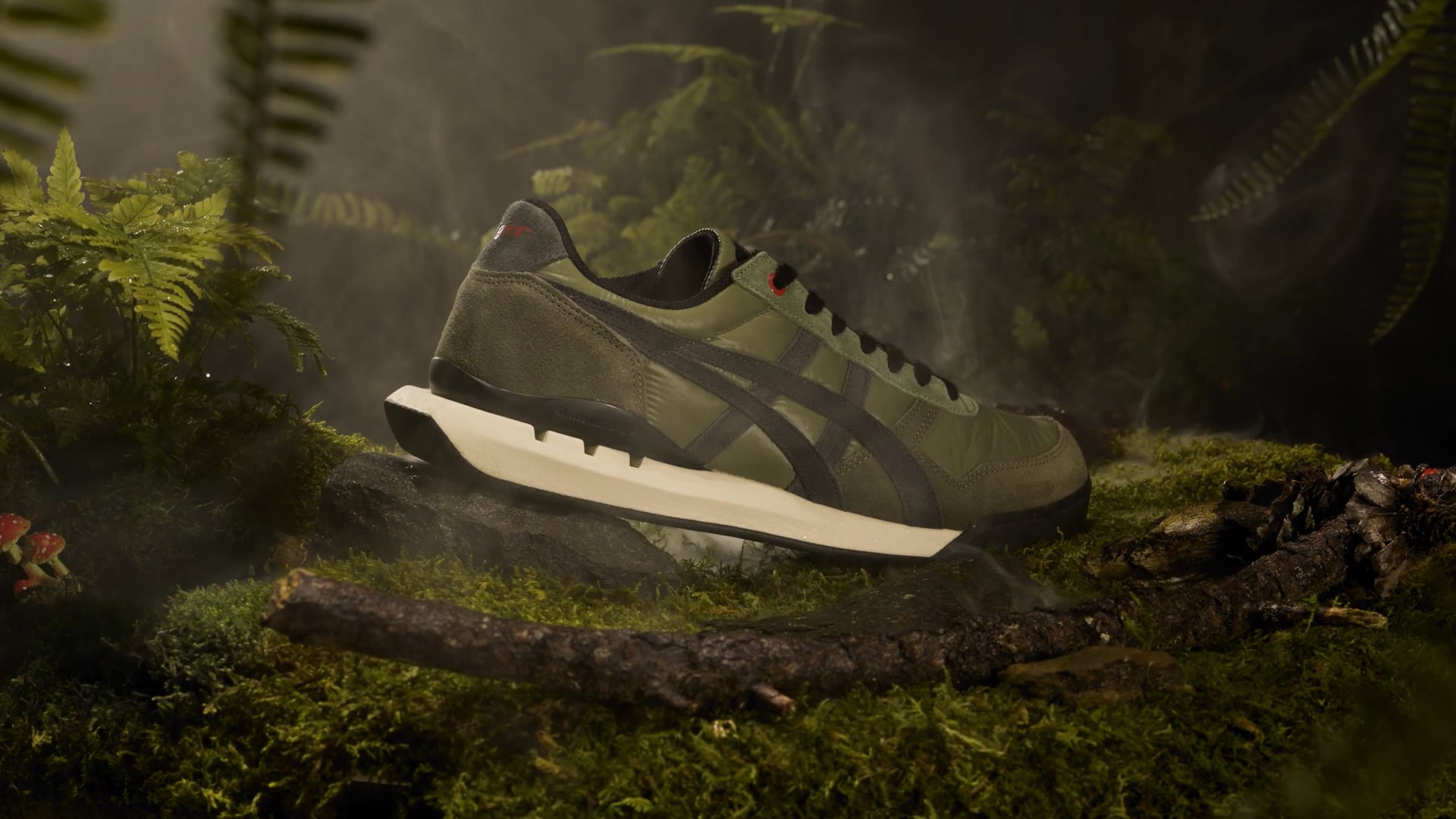 video of an Onitsuka Tiger Ultimate 81 sneaker in mossy green colors with red details shot in a misty forest floor scene with ferns, moss, mushrooms, and other detritus.  There is still dew on the leaves and the gentle sounds of a stream and the forest waking up play in the background